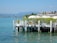 A wooden deck extends over a serene body of water, adorned with white railings and flowering plants. Several large umbrellas provide shade over lounge chairs. The backdrop features a distant view of rolling hills and coastline under a clear blue sky.