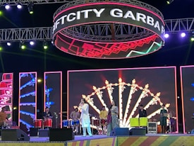 A vibrant stage with multiple musicians performing. Colorful lights and a large LED display create a festive atmosphere. The performers are dressed in traditional attire and playing various drums. Above the stage, a circular sign displays the words 'GIFT CITY GARBA N.'