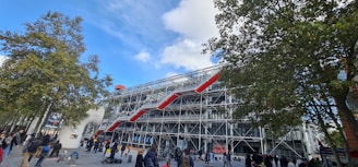 An urban scene featuring a large, modern building with a striking architectural design characterized by its visible structural framework and bright red external escalators. Trees are present in the foreground, with people walking or gathering in the plaza area.