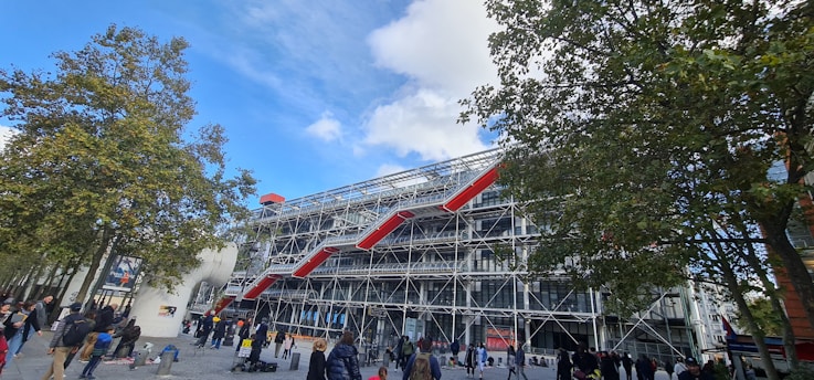 An urban scene featuring a large, modern building with a striking architectural design characterized by its visible structural framework and bright red external escalators. Trees are present in the foreground, with people walking or gathering in the plaza area.