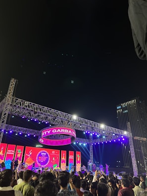 A lively outdoor event with a large crowd gathered in front of a brightly lit stage. The stage features an array of colorful lights and banners, including a prominent circular sign displaying 'CITY GARBA'. The audience is animated, with many people capturing the moment on their phones or clapping along. Tall buildings are visible in the background, hinting at an urban setting. Musicians and performers are positioned on the stage, contributing to the festive atmosphere.