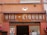 A vintage shop sign reads 'VINI - LIQUORI' with the names 'GIOVANNI' and 'ZAMPIERI' underneath. The sign is set against a building with an orange facade, adorned with some wires and a small wall-mounted light fixture. There are decorative red wreaths on either side of a glass doorway below the sign.