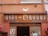 A vintage shop sign reads 'VINI - LIQUORI' with the names 'GIOVANNI' and 'ZAMPIERI' underneath. The sign is set against a building with an orange facade, adorned with some wires and a small wall-mounted light fixture. There are decorative red wreaths on either side of a glass doorway below the sign.