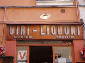 A vintage shop sign reads 'VINI - LIQUORI' with the names 'GIOVANNI' and 'ZAMPIERI' underneath. The sign is set against a building with an orange facade, adorned with some wires and a small wall-mounted light fixture. There are decorative red wreaths on either side of a glass doorway below the sign.
