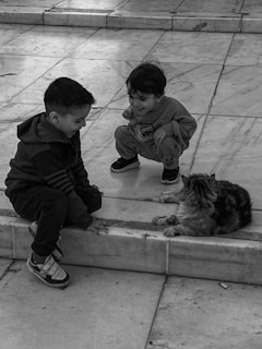 Two young children are interacting with a cat on a tiled surface. One child is sitting on the edge while the other is squatting, both focusing their attention on the cat lying in front of them.