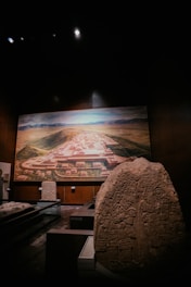 A dimly lit museum exhibit features a large, carved stone with ancient inscriptions and figures prominently displayed in the foreground. In the background, a detailed mural or model depicts an ancient city layout, surrounded by a landscape of mountains and valleys.