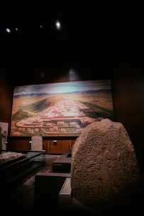 A dimly lit museum exhibit features a large, carved stone with ancient inscriptions and figures prominently displayed in the foreground. In the background, a detailed mural or model depicts an ancient city layout, surrounded by a landscape of mountains and valleys.