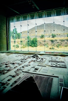 A detailed architectural model is displayed in the foreground, representing a complex of ancient structures and layout. Large windows in the background reveal a pyramid and natural landscape with trees and shrubs, bathed in daylight.