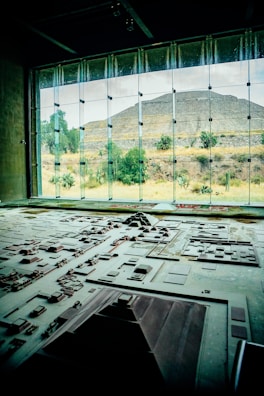 A detailed architectural model is displayed in the foreground, representing a complex of ancient structures and layout. Large windows in the background reveal a pyramid and natural landscape with trees and shrubs, bathed in daylight.