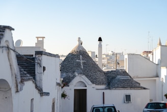 A quaint street scene featuring traditional houses with unique conical, stone roofs. The foreground includes a small, narrow street with a parked car and flowers hanging by the entrance of one house. The background shows more buildings with similar architecture, set against a clear blue sky.