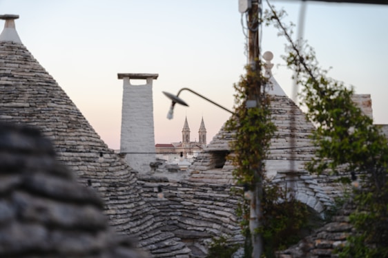 A close-up view of stone cone-shaped rooftops, characteristic of traditional trulli homes, with a background featuring a church steeple. The image has a soft, pastel-colored sky at dusk. Vines and a streetlight are also visible, adding texture and depth to the architectural scene.