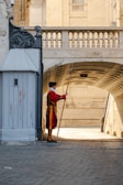 A guard dressed in colorful traditional attire stands at attention beside a gray guardhouse, holding a long pole. The setting includes a cobblestone pathway that leads under an elegant stone archway, framed by historical architecture in warm tones.