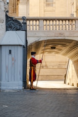A guard dressed in colorful traditional attire stands at attention beside a gray guardhouse, holding a long pole. The setting includes a cobblestone pathway that leads under an elegant stone archway, framed by historical architecture in warm tones.