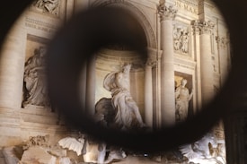 A classical stone sculpture surrounded by ornate architectural details, such as columns and decorative carvings. The scene is viewed through a circular frame, adding depth. The sculptures depict historical or mythological figures, characterized by flowing robes and intricate craftsmanship.