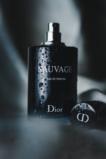 A dark, elegant perfume bottle stands upright, adorned with water droplets. The bottle features the words 'Sauvage' and 'Dior' in sleek, reflective lettering. A blurred, moody background adds to the sophisticated and luxurious vibe.