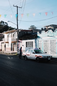 A street scene depicts a taxi parked alongside colorful buildings with intricate ironwork. Above, a series of multicolored triangular flags are strung across. A person is walking near the taxi.