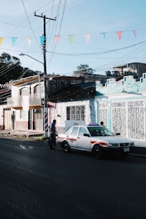 A street scene depicts a taxi parked alongside colorful buildings with intricate ironwork. Above, a series of multicolored triangular flags are strung across. A person is walking near the taxi.