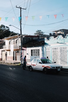 A street scene depicts a taxi parked alongside colorful buildings with intricate ironwork. Above, a series of multicolored triangular flags are strung across. A person is walking near the taxi.