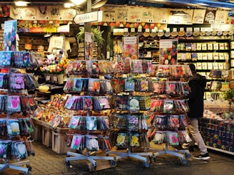 A market stall is filled with various packet seeds on display racks. The packets feature colorful images of flowers and plants. A person observes the packets while another person is visible in the background attending the stall. The environment appears busy and vibrant with a wide range of gardening supplies.