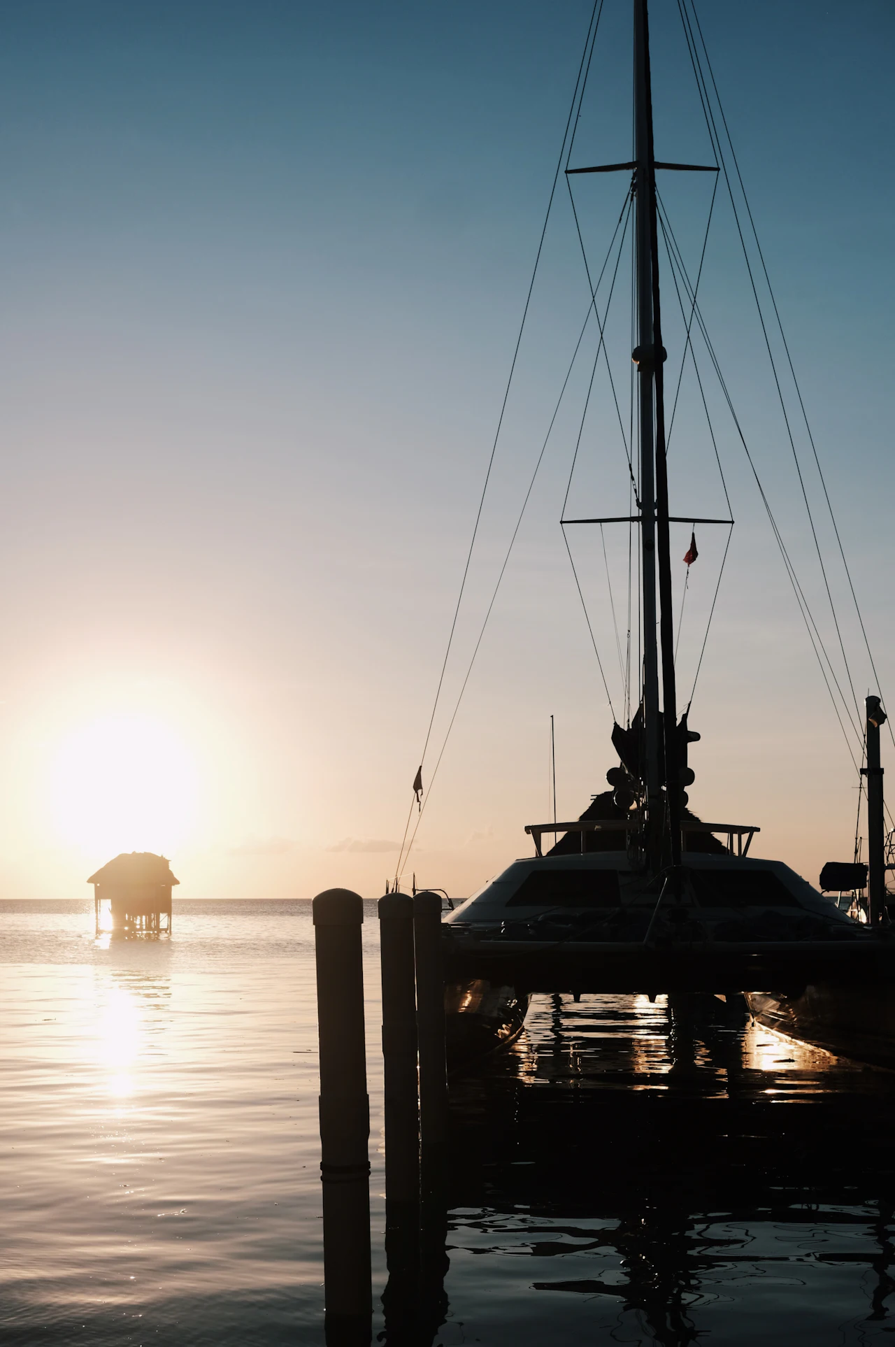 A sailing boat silhouetted against a low sun over calm water, with an over-water hut on the horizon.