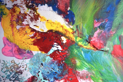 A vibrant abstract painting featuring a blend of vivid colors including red, yellow, blue, green, and pink. The texture is rich, with visible brushstrokes and layers of paint creating a dynamic and energetic composition.