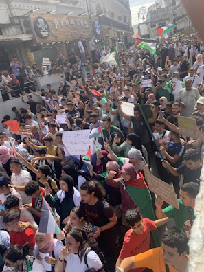 A large crowd is gathered for a protest, with many people holding flags and signs. The scene is lively, with participants raising their arms and displaying various banners. The area appears to be a busy urban street with buildings in the background.