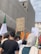 A group of people participate in a protest, holding various signs and flags. One prominent sign reads 'Killing children is a war crime,' and features a small flag. The atmosphere is one of activism, with individuals standing close together. The background shows a building under construction or renovation.