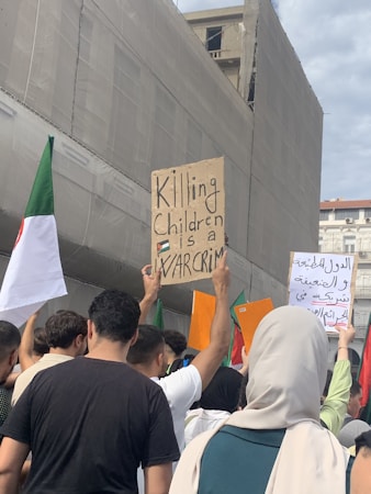 A group of people participate in a protest, holding various signs and flags. One prominent sign reads 'Killing children is a war crime,' and features a small flag. The atmosphere is one of activism, with individuals standing close together. The background shows a building under construction or renovation.