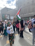 A group of people gathers in an urban setting, holding various flags, including Palestinian and Algerian flags. One child is prominently raising a Palestinian flag. The crowd includes individuals wearing headscarves and colorful clothing. A large screen in the background displays the Algerian flag.