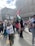 A group of people gathers in an urban setting, holding various flags, including Palestinian and Algerian flags. One child is prominently raising a Palestinian flag. The crowd includes individuals wearing headscarves and colorful clothing. A large screen in the background displays the Algerian flag.