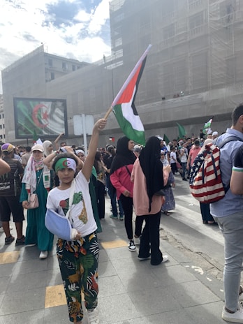 A group of people gathers in an urban setting, holding various flags, including Palestinian and Algerian flags. One child is prominently raising a Palestinian flag. The crowd includes individuals wearing headscarves and colorful clothing. A large screen in the background displays the Algerian flag.