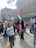A group of people gathers in an urban setting, holding various flags, including Palestinian and Algerian flags. One child is prominently raising a Palestinian flag. The crowd includes individuals wearing headscarves and colorful clothing. A large screen in the background displays the Algerian flag.