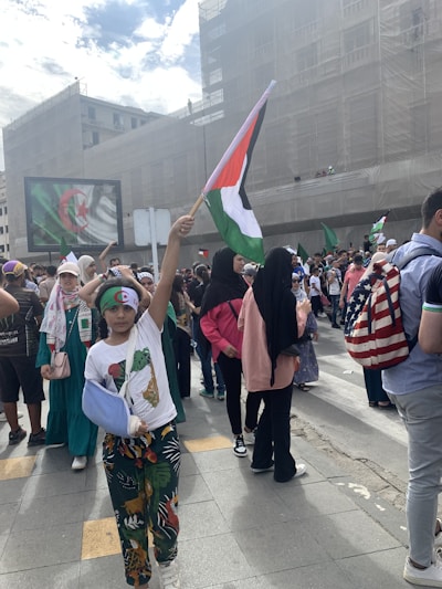 A group of people gathers in an urban setting, holding various flags, including Palestinian and Algerian flags. One child is prominently raising a Palestinian flag. The crowd includes individuals wearing headscarves and colorful clothing. A large screen in the background displays the Algerian flag.