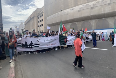 A large group of people is participating in a protest, holding banners and flags. The banner displays messages in Arabic and English, including 'No to killing children, women, and civilians.' The scene is set on a street beside a large building under renovation. Participants appear engaged and are waving various flags.