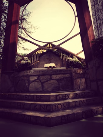 An outdoor altar is framed by a large circular architectural structure with wooden beams. The altar features a stone surface with engraved text from the Lord's Prayer. Candles and floral arrangements adorn the top of the altar. The background shows trees and part of a stone wall, with sunlight casting a warm glow.