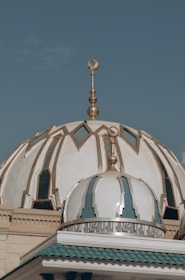 An ornate mosque dome with intricate designs and patterns, featuring gold decorative elements and Arabic calligraphy. The dome is set against a clear blue sky.