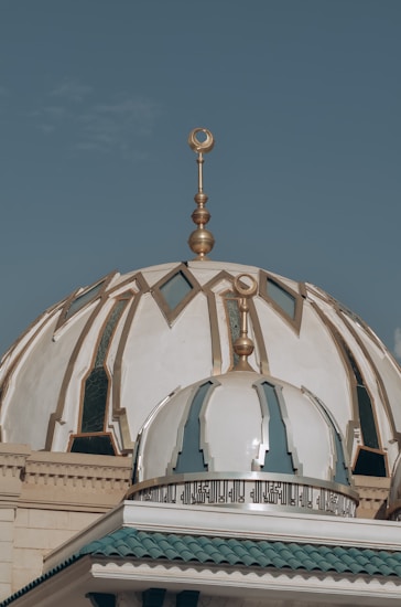 An ornate mosque dome with intricate designs and patterns, featuring gold decorative elements and Arabic calligraphy. The dome is set against a clear blue sky.