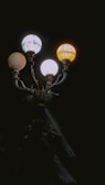 A vintage street lamp with three ornate globes casting yellow and white light. The intricate metalwork is visible in the faint light against a dark background.