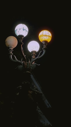 A vintage street lamp with three ornate globes casting yellow and white light. The intricate metalwork is visible in the faint light against a dark background.