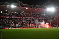 A football stadium packed with enthusiastic fans displaying numerous flags and banners. The crowd is vibrant and energetic, with some areas illuminated by red flares. The stadium is covered, and many people are holding lights, adding to the dynamic atmosphere.