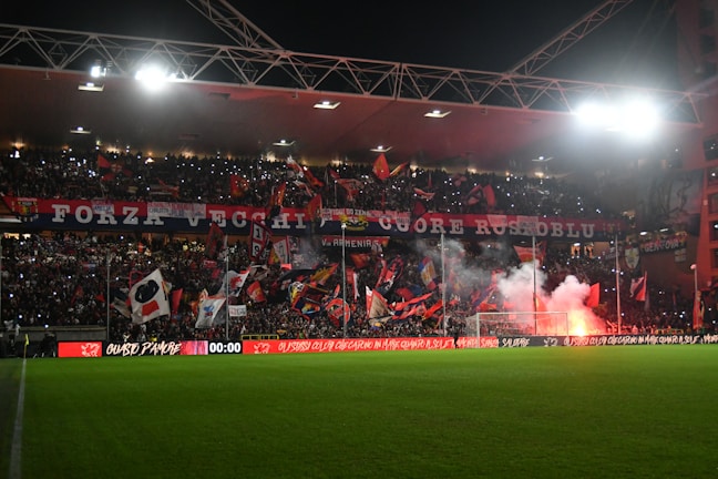 A football stadium packed with enthusiastic fans displaying numerous flags and banners. The crowd is vibrant and energetic, with some areas illuminated by red flares. The stadium is covered, and many people are holding lights, adding to the dynamic atmosphere.