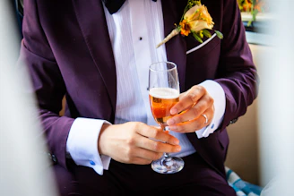 A person is elegantly dressed in a dark purple tuxedo with a white shirt and a bow tie. They are holding a glass of amber-colored beverage, possibly champagne or sparkling wine. A boutonniere with a yellow rose is attached to the lapel.