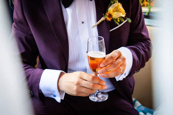 A person is elegantly dressed in a dark purple tuxedo with a white shirt and a bow tie. They are holding a glass of amber-colored beverage, possibly champagne or sparkling wine. A boutonniere with a yellow rose is attached to the lapel.
