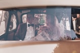 A wedding couple is seated inside a vintage car. The groom is dressed in a dark suit with a bow tie, and the bride wears a veil and holds a bouquet of flowers. Outside, there are people gathered, possibly guests or onlookers.