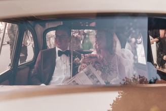 A wedding couple is seated inside a vintage car. The groom is dressed in a dark suit with a bow tie, and the bride wears a veil and holds a bouquet of flowers. Outside, there are people gathered, possibly guests or onlookers.