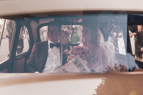 A wedding couple is seated inside a vintage car. The groom is dressed in a dark suit with a bow tie, and the bride wears a veil and holds a bouquet of flowers. Outside, there are people gathered, possibly guests or onlookers.