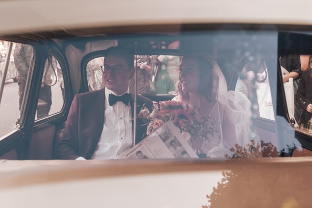 A wedding couple is seated inside a vintage car. The groom is dressed in a dark suit with a bow tie, and the bride wears a veil and holds a bouquet of flowers. Outside, there are people gathered, possibly guests or onlookers.