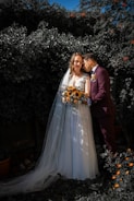 A couple in wedding attire poses affectionately in a garden setting. The bride wears a white gown with a long veil and holds a bouquet of sunflowers, while the groom dons a dark suit with a boutonniere. They are surrounded by lush greenery, and there are small orange flowers in the foreground.