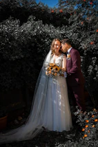 A couple in wedding attire poses affectionately in a garden setting. The bride wears a white gown with a long veil and holds a bouquet of sunflowers, while the groom dons a dark suit with a boutonniere. They are surrounded by lush greenery, and there are small orange flowers in the foreground.