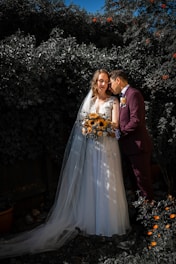 A couple in wedding attire poses affectionately in a garden setting. The bride wears a white gown with a long veil and holds a bouquet of sunflowers, while the groom dons a dark suit with a boutonniere. They are surrounded by lush greenery, and there are small orange flowers in the foreground.